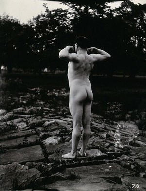 view A man posing naked, with his back to the camera, standing on a rocky outcrop.