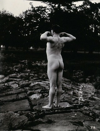 A man posing naked, with his back to the camera, standing on a rocky outcrop.