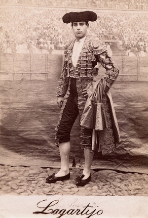 view A bullfighter, Rafael Molina Sanchez "Lagartijo", posing in front of a painted backdrop of a bullring crowd, in a cobbled courtyard. Photograph, ca.1890/1910.