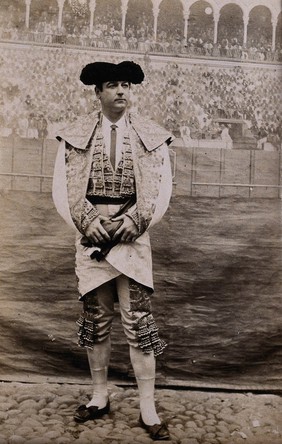 A bullfighter, posing in front of a painted backdrop of a bullring, in a cobbled courtyard. Photograph, first quarter of the twentieth century.