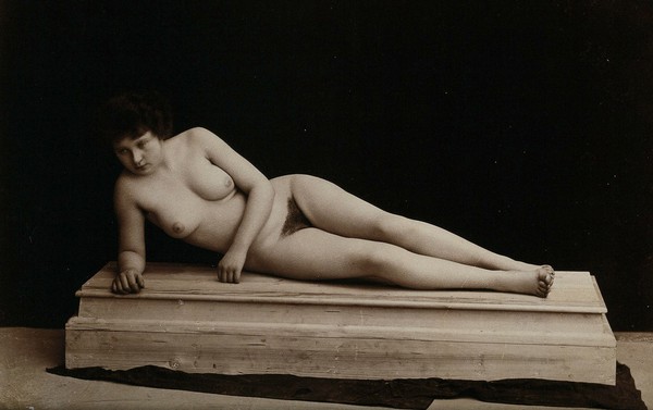 A young woman posing naked in a photographic studio, lying on a wooden plinth. Photograph, ca.1900.