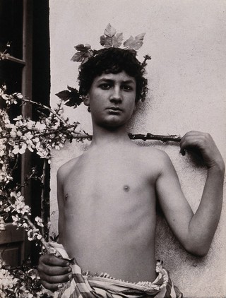 A Sicilian boy, naked from the waist up, his hand level with his shoulder, crowned with leaves. Photograph by W. von Gloeden, 1901.