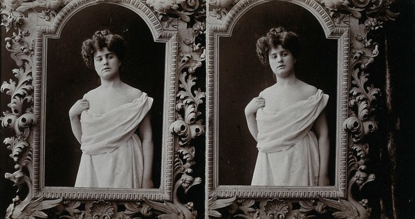 A woman posing wrapped in a sheet, looking through a screen giving the effect of the frame of a looking-glass, in a photographic studio.