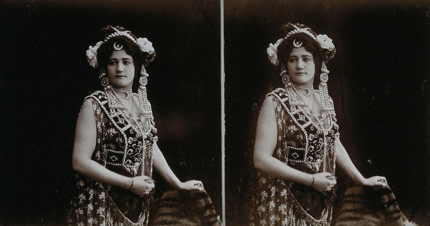 A young woman posing in a photographic studio, wearing an unusual dress, in front of a plain backdrop.