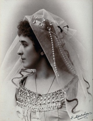 A woman posing in a photographic studio, wearing a bridal? dress and veil, in front of a plain backdrop.