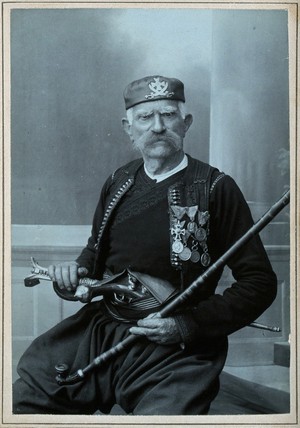 view A Montenegrin man wearing national dress with medals, with a pistol in his belt and holding a tobacco pipe.