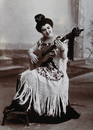 view A seated woman, playing a guitar, wearing a fringed shawl. Photograph, ca.1900.