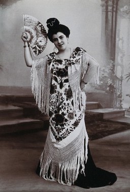 A standing woman, holding aloft a fan, wearing a fringed shawl, in front of a painted backdrop in a photographic studio. Photograph, ca.1900.