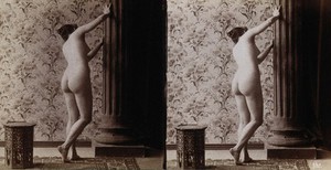 view A young woman posing naked in a photographic studio, with her back to the camera looking round a column, a small Moorish table behind her. Stereo photograph, ca.1900.