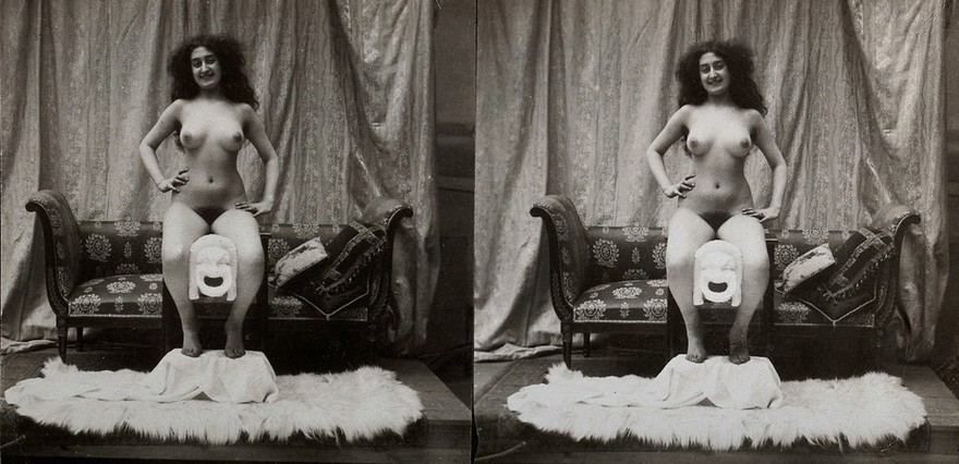 A young woman, full-length, posed naked sitting on a table with her feet on a fur-covered stool, in a photographic studio. Photograph, ca.1900.