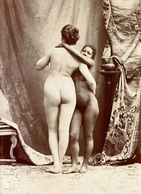 Two women posing naked in a photographic studio, standing embracing each other in front of some studio props.