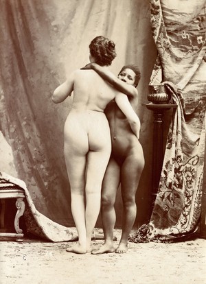 view Two women posing naked in a photographic studio, standing embracing each other in front of some studio props.