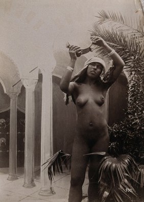 A woman posing naked in a photographic studio, holding aloft a tambourine, standing in front of a painted backdrop. Photograph, c.1900.