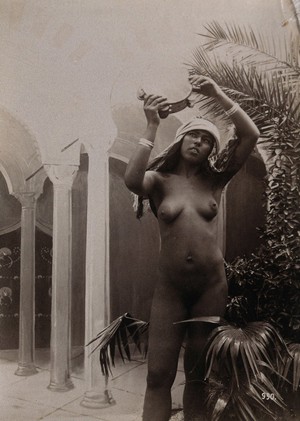 view A woman posing naked in a photographic studio, holding aloft a tambourine, standing in front of a painted backdrop. Photograph, c.1900.