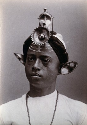 A young Indian man wearing an elaborate headress and makeup, in a studio setting. Photograph, ca.1900.