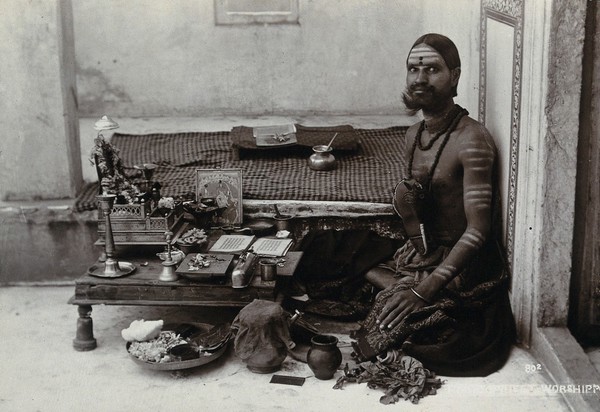 An Indian man wearing elaborate jewellery and make-up, squatting in front of a small table on which is a shrine, perhaps in a temple. Photograph, ca.1900.