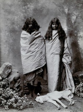 A man and woman of the Toda tribe standing in a photographic studio, with a dog lying at their feet.