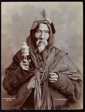 A bearded elderly man, carrying a prayer wheel and a rosary, in a studio setting.