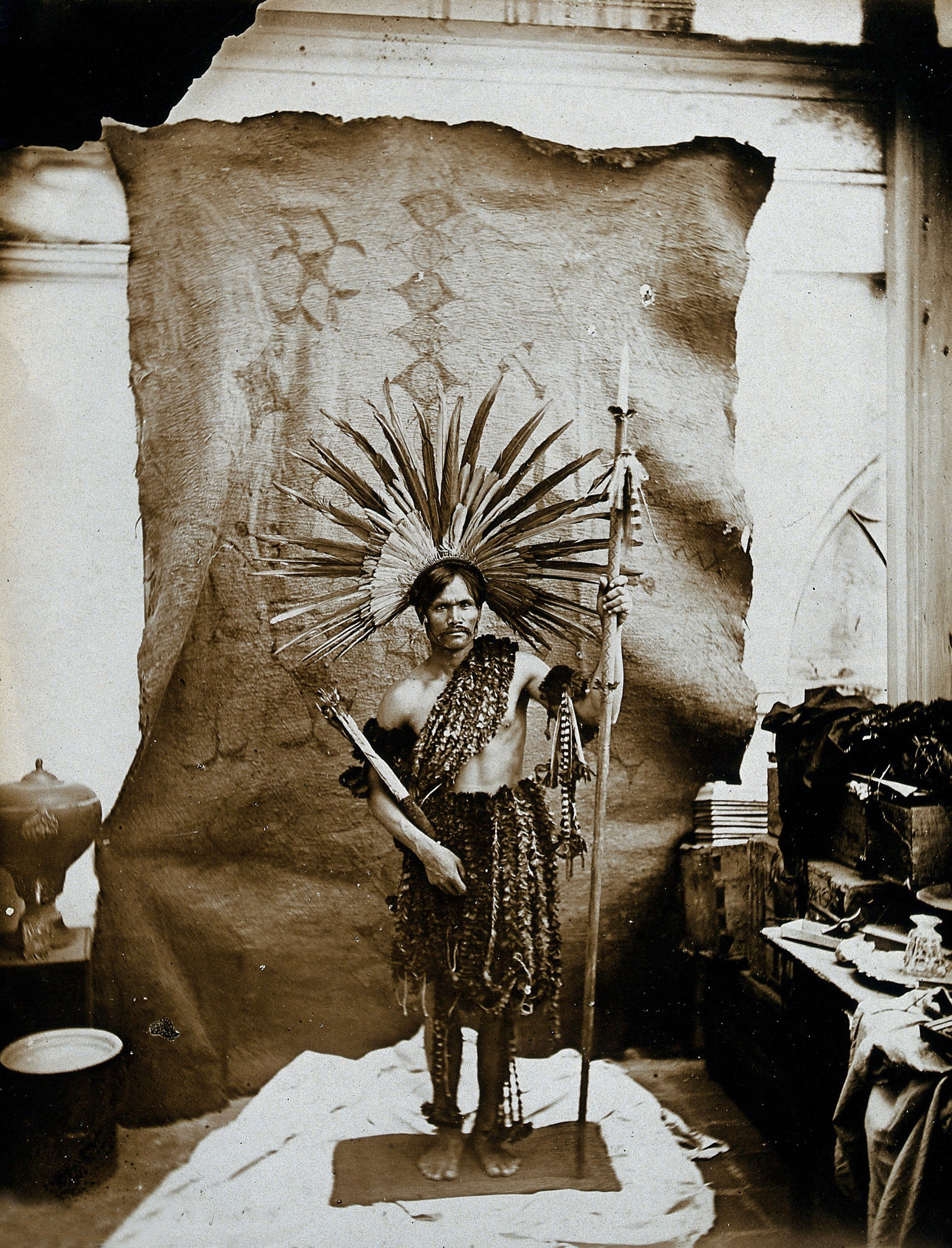 A man of an Amazonian tribe, holding a spear and wearing a feathered head-dress, in a photographic studio. 