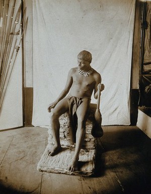 view A man posing on a pedestal, holding a club (?), in a photographic studio.