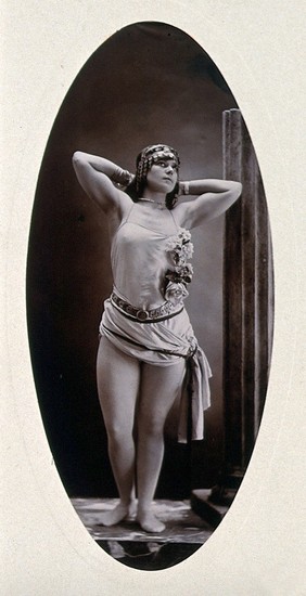 A young woman posing standing on a podium beside a truncated classical column in a photographic studio. Photograph by Portland Studio.