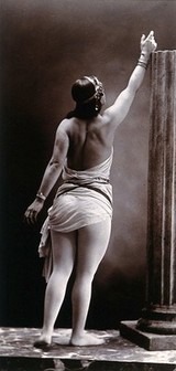 A young woman posing semi-naked, back view, standing on a podium beside a truncated classical column, in a photographic studio. Photograph by Portland Studio.