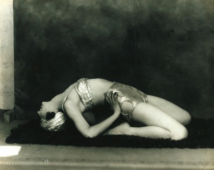 Marguerite Agniel posing leaning back wearing a two-piece costume and matching turban, in a photographic studio.