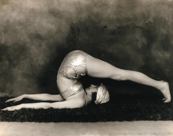 Marguerite Agniel posing with her back arched and legs stretched out over her head, wearing a two-piece costume and matching turban, in a photographic studio.