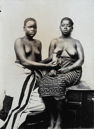 Two women from Zanzibar, one milking the other's right breast into a cup.