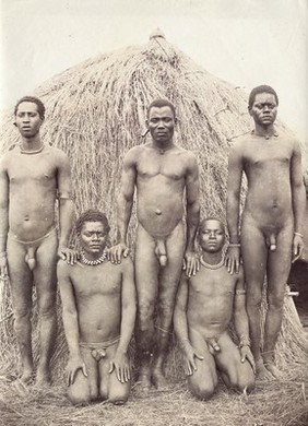 Five nude African men, in front of a hut.