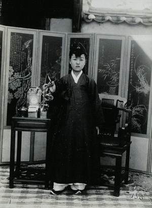 view A young woman wearing a plain robe and a tasselled hat, standing in front of a painted screen.