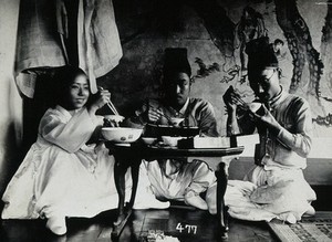 view A group of people eating a meal, seated behind a small table, in front of a painted screen.