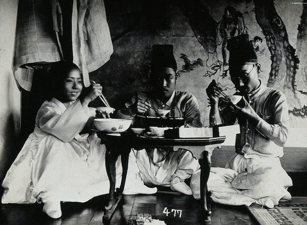 A group of people eating a meal, seated behind a small table, in front of a painted screen.