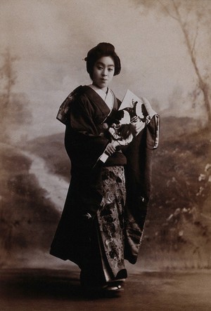 view A young woman posing in a photographic studio, holding a fan, in front of a painted backdrop of a rural scene.