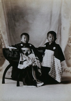 view Two boys in a photographic studio, one seated on a chair, in front of a curtained backdrop.