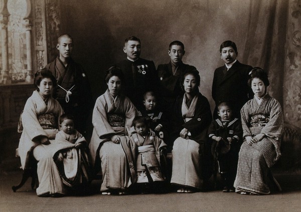 A large family group in a photographic studio, the women seated, in front of a painted backdrop.