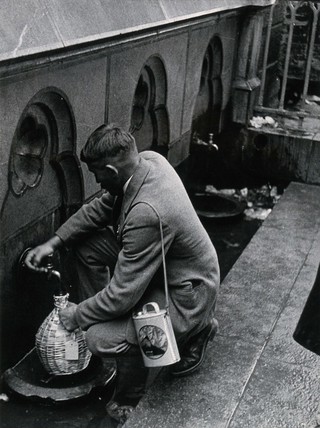 Lourdes, France: a pilgrim fills a flask with sacred spring water from a tap. Photograph, ca. 1937.