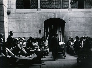 view Lourdes, France: an invalid being wheeled through to the baths while other ill pilgrims await their turn for immersion by the priest. Photograph, ca. 1937.