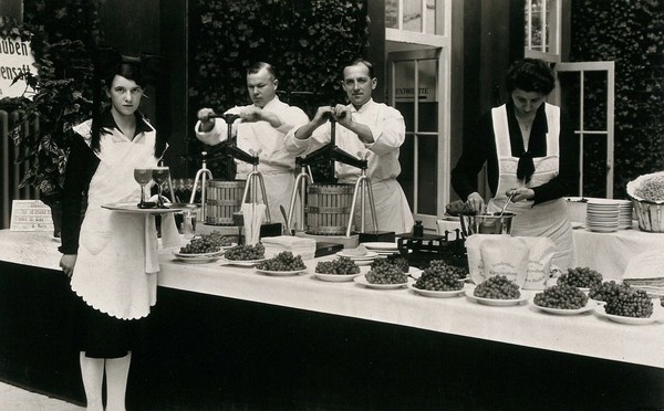 Baden-Baden, Germany: waiters in a grape-cure establishment standing next to a buffet with plates of grapes. Photographic postcard, ca. 1930.