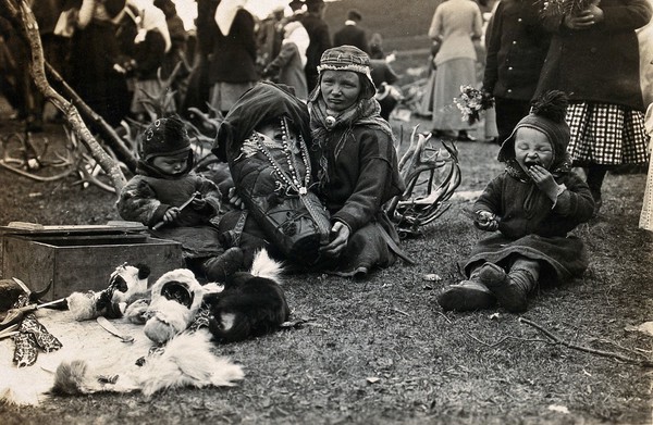 A Lapp (Sami) woman holding her baby in a child carrier, her two toddlers beside her. Photograph by Davey & Hackney, ca. 1913.