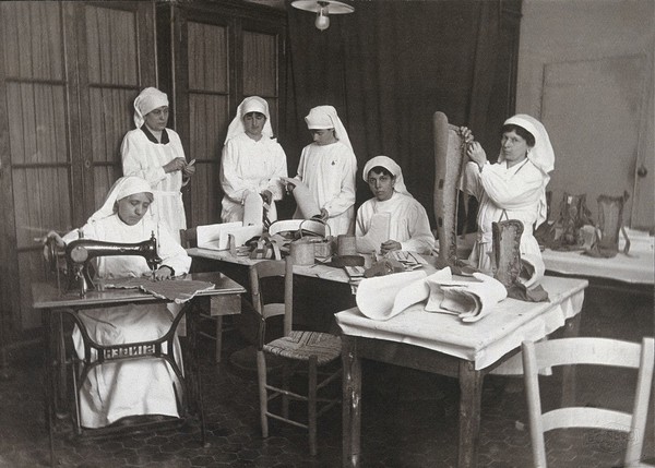 Artificial limb factory in Rome: six women working at benches, one using a sewing machine and one stitching the back of a full-length leg. Photograph, 1914/1918.