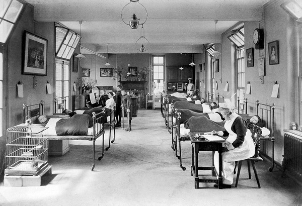 Alexandra Hospital, East Clandon: a ward in which some children are in bed while others stand around a piano being played by a nurse. Photograph, 1913(?).