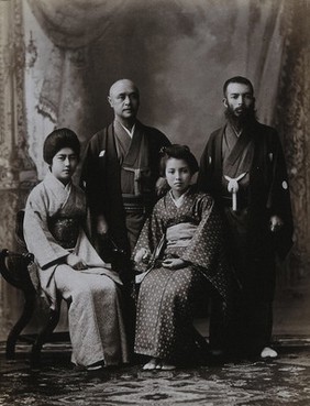 A family group in a photographic studio, the women seated, in front of a curtained backdrop.