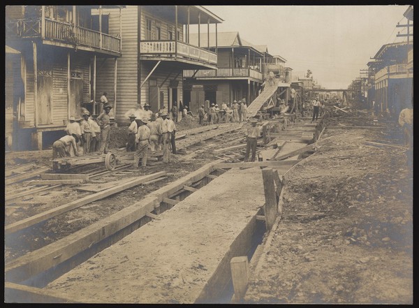 Colón, Panama: men construct sewers in the middle of a road lined with wooden houses, as part of a programme of sanitary work implemented during the construction of the Panama Canal. Photograph, 1910.