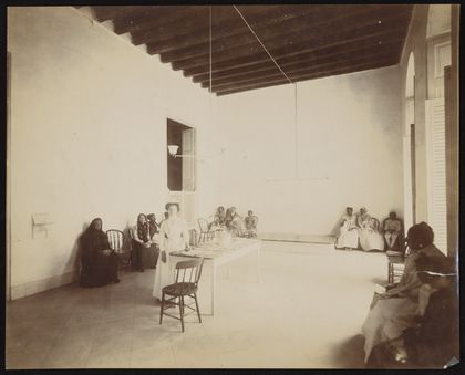 Waiting room in a dispensary for the treatment of tuberculosis, Cuba: a nurse stands beside the reception table, surrounded by seated patients. Photograph, 1902.
