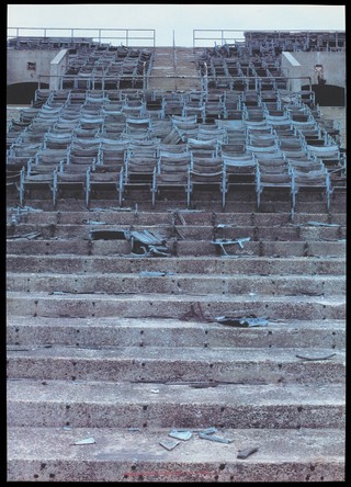 Concrete steps within a stadium with broken chairs representing the destructive effect of AIDS. Colour silk screen print after Dennis Adams, 1993.