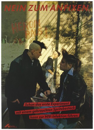 A man with a skin head and leather jacket talks to a woman with a skin head against a metal fence with grafitti on a wall in the background: "Heroin Raus!" [heroin out]; a warning about the dangers of sharing needles and contracting AIDS by the Deutsche AIDS-Hilfe e.V. Colour lithograph by R. Warzecha, M. Jahreiss and D. Pusch.