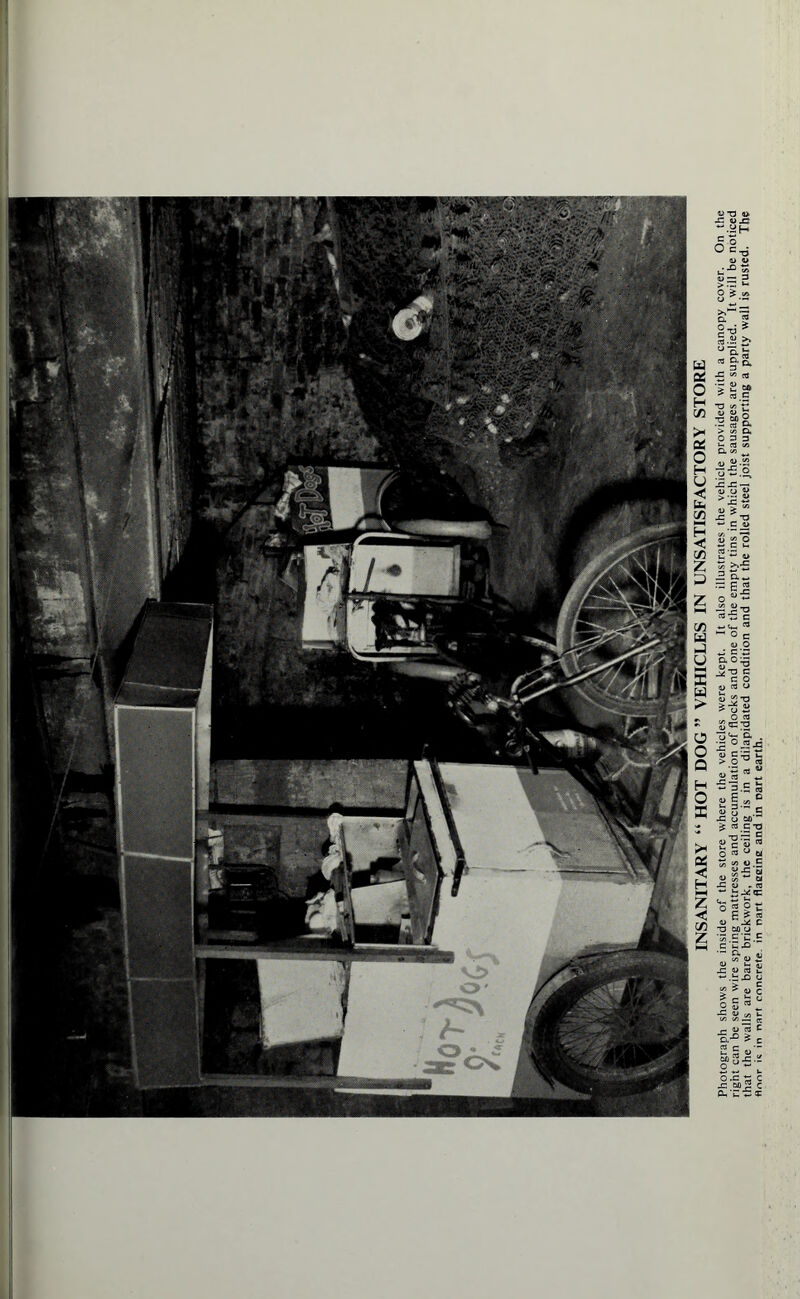 INSANITARY “ HOT DOG ” VEHICLES IN UNSATISFACTORY STORE Photograph shows the inside of the store where the vehicles were kept. It also illustrates the vehicle provided with a canopy coyer. On the right can be seen wire spring mattresses and accumulation of flocks and one of the empty tins in which the sausages are supplied, ft will be noticed that the walls are bare brickwork, the ceiling is in a dilapidated condition and that the rolled steel joist supporting a party wall is rusted. The floor is in nart concrete, in nart flaeeine and in part earth.