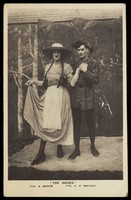 view Two members of a military concert party pose on stage: one is in drag with a large hat and pigtails. Photographic postcard, 1915-1916.