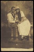 view Two sailors, one in drag, embracing while sitting on a stage balcony. Photographic postcard, 191-.